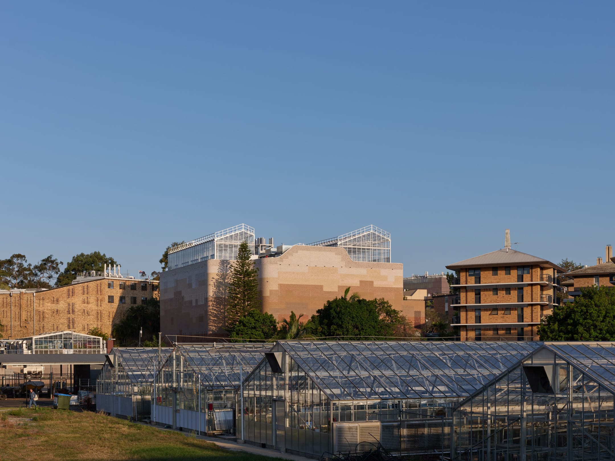 Modern brick buildings with glass greenhouses in foreground under clear blue sky, urban garden setting.