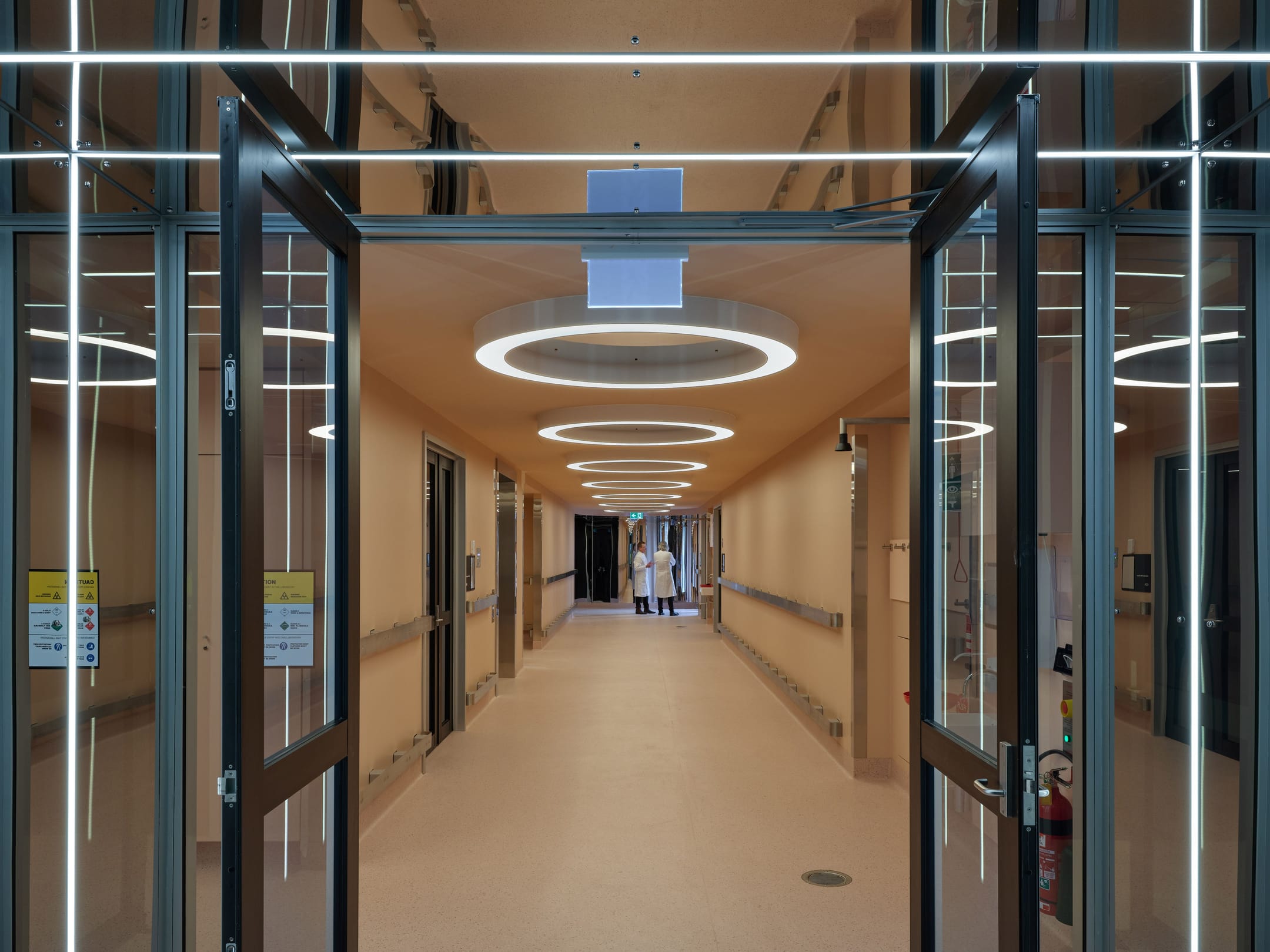 Modern hospital corridor with circular LED ceiling lights, glass doors with safety information, and two people walking in the distance.
