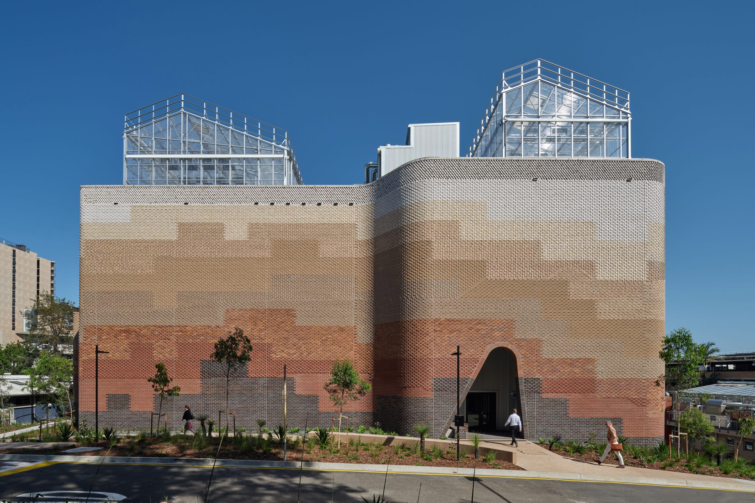 Modern brick building with curved arched entrance, glass greenhouses on roof, clear blue sky, landscaping with young trees.