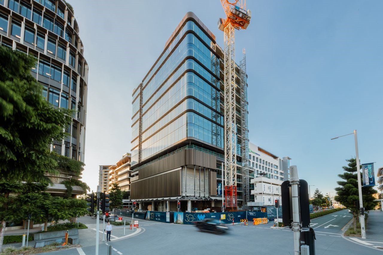Modern construction site with glass and bronze-toned building under development, surrounded by residential apartments and construction cranes.