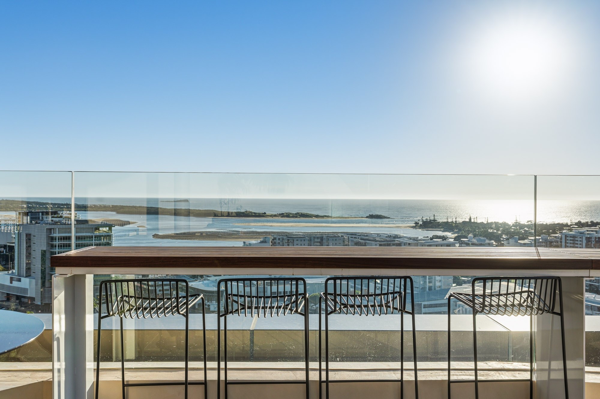 Modern rooftop bar with four black metal stools overlooking a coastal bay and waterfront cityscape on a clear sunny day
