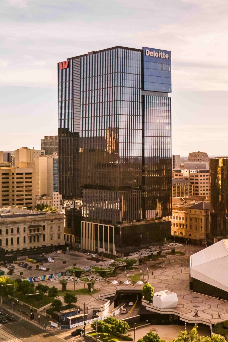 Deloitte office tower with vertical ribbed facade in downtown cityscape at sunset, surrounded by other buildings and plaza below.