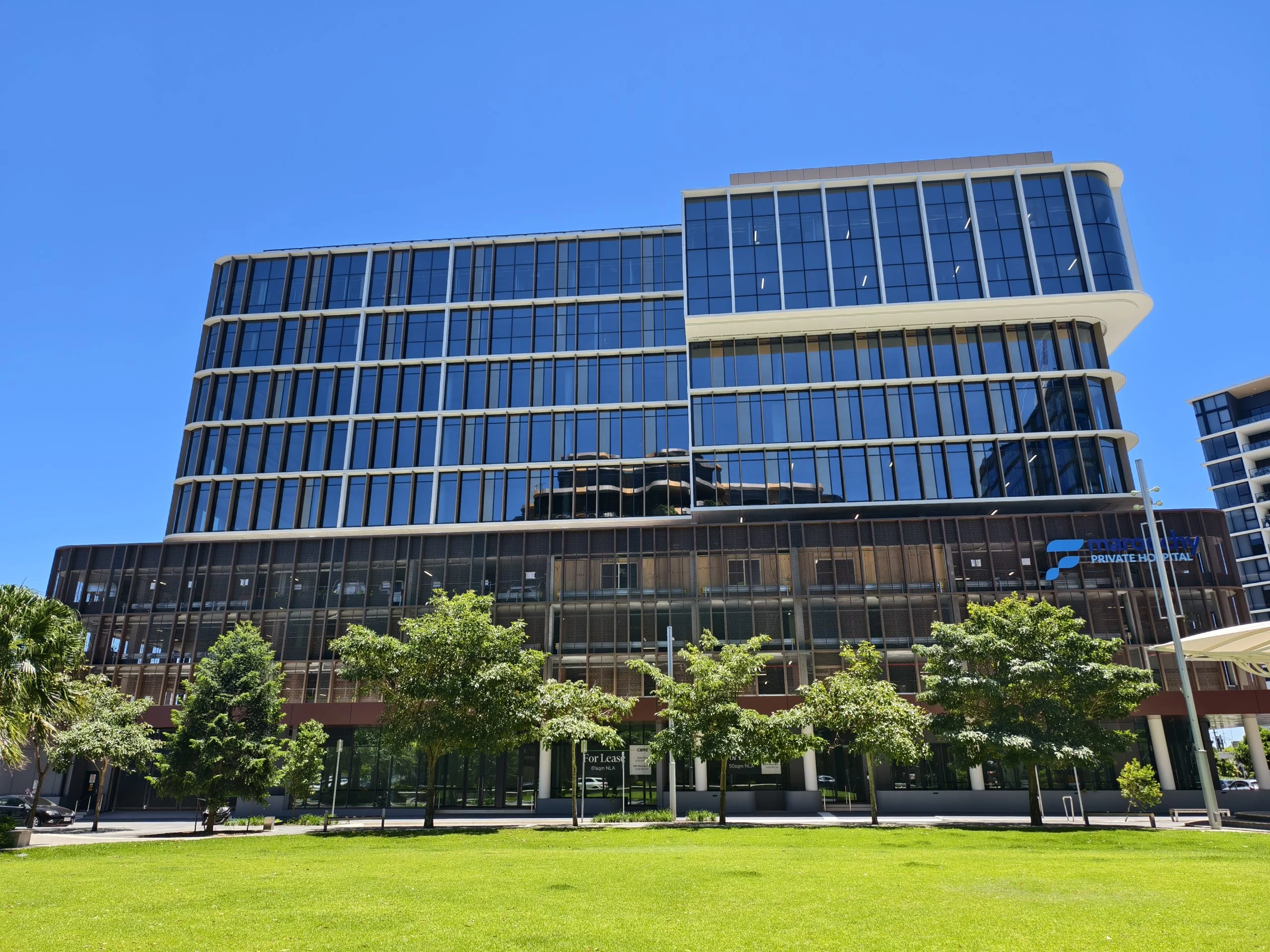 Modern multi-story office building with curved glass facade, dark lower levels, and green plaza with trees in foreground under clear blue sky.