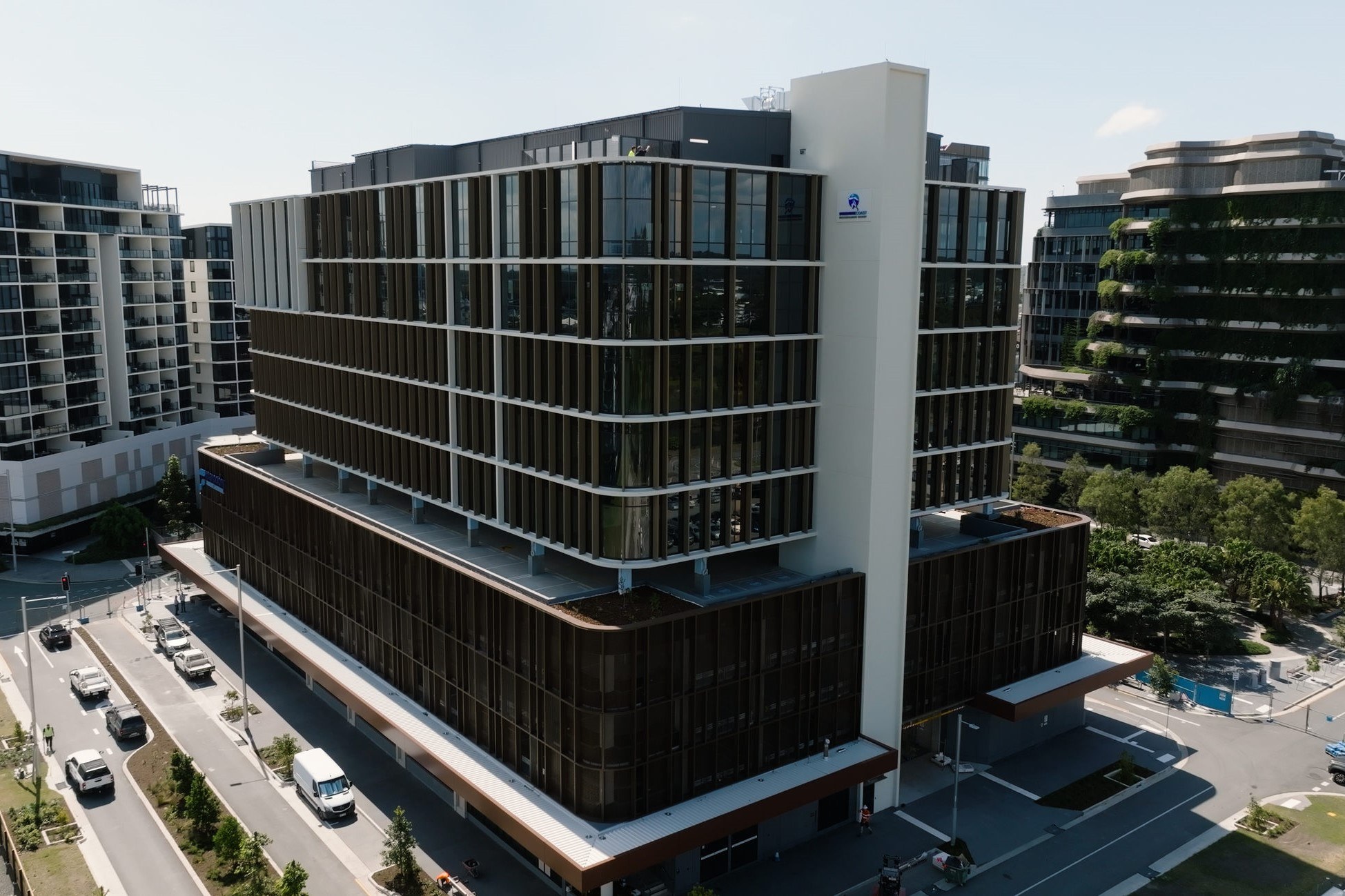 Modern curved office building with dark vertical slats and white tower, surrounded by residential apartments and street-level parking.
