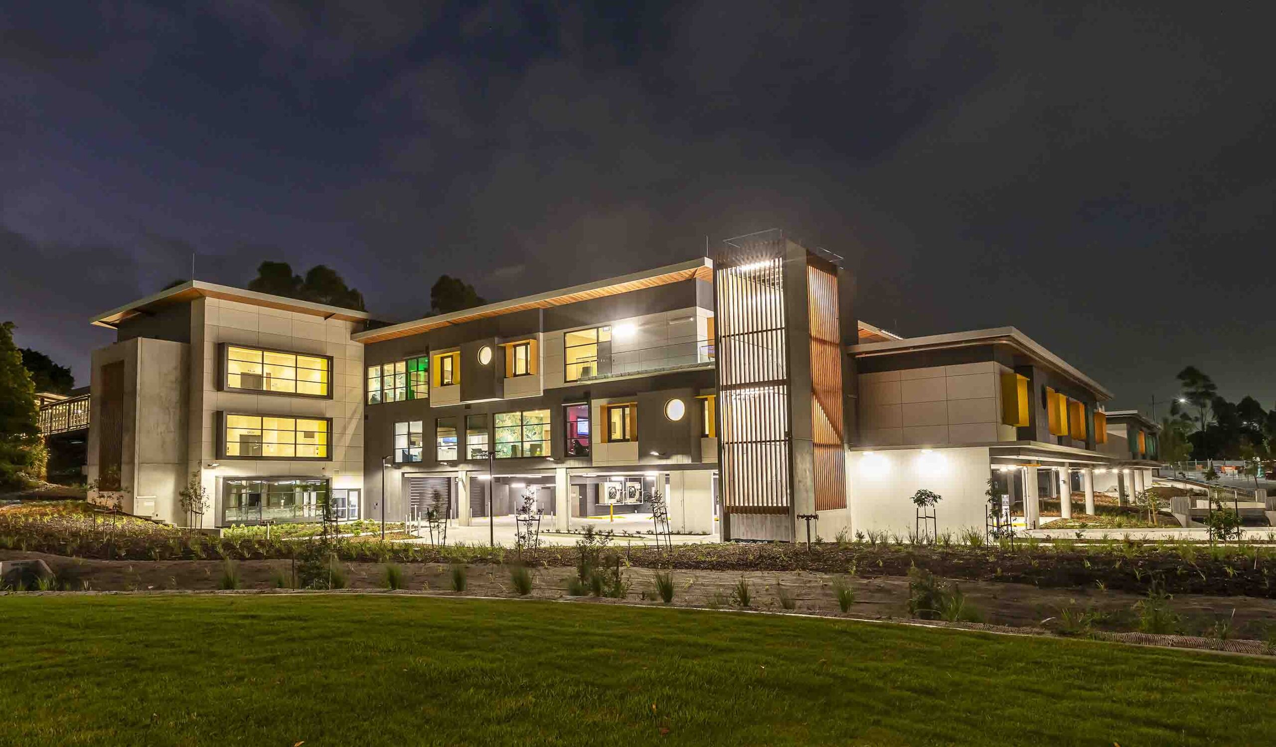 Modern multi-story building with illuminated windows, concrete and metal facade, landscaping, and green lawn at dusk.