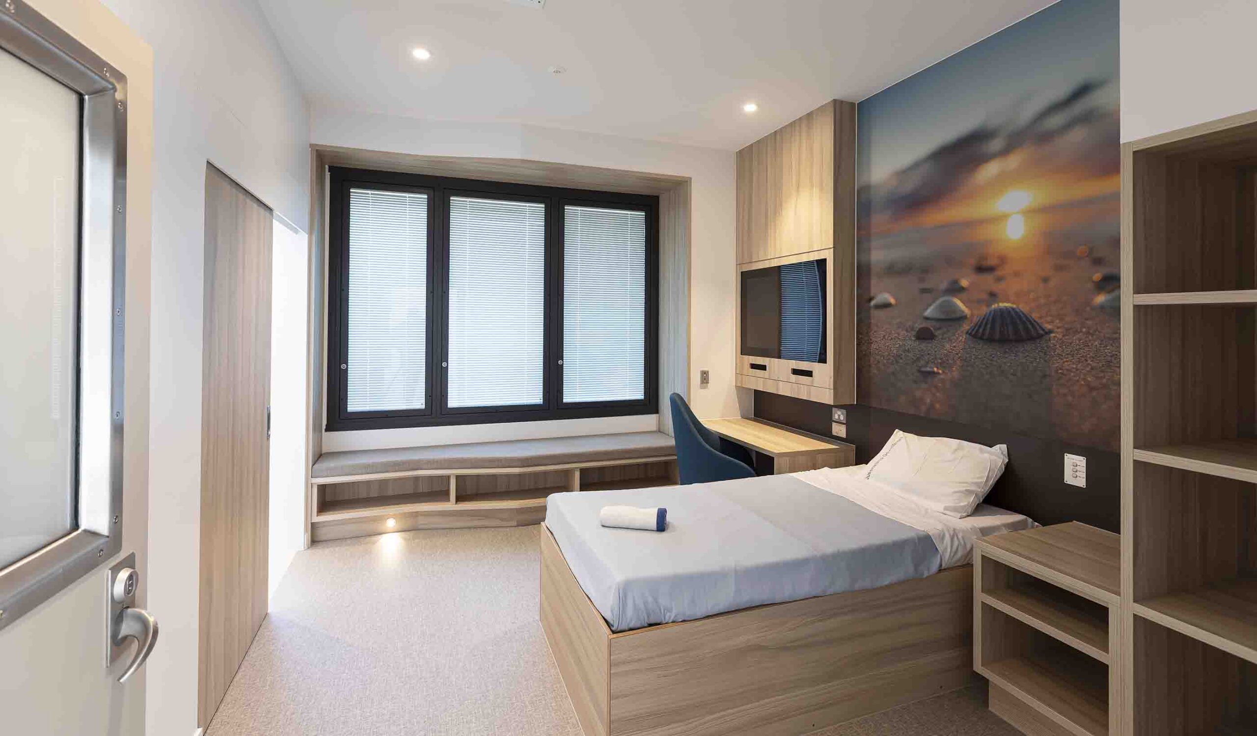 Modern bedroom with bed, desk, window seat, shelving, and desert landscape artwork on dark accent wall.