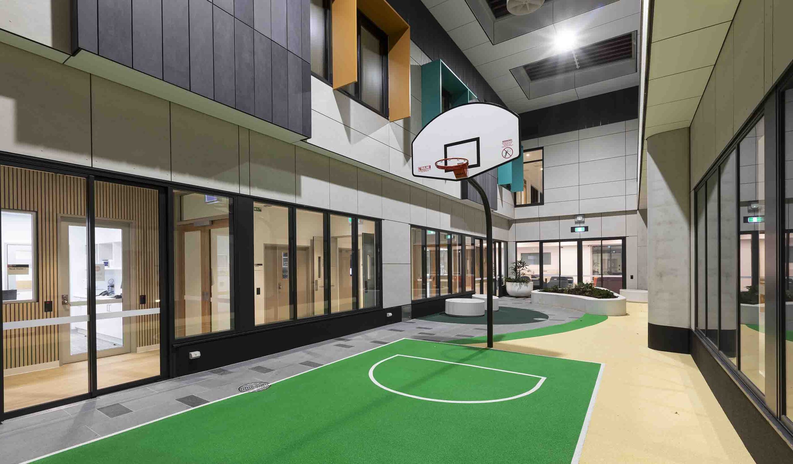 Modern indoor sports facility with basketball court, hoop, green playing surface, and glass-walled rooms along the sides.