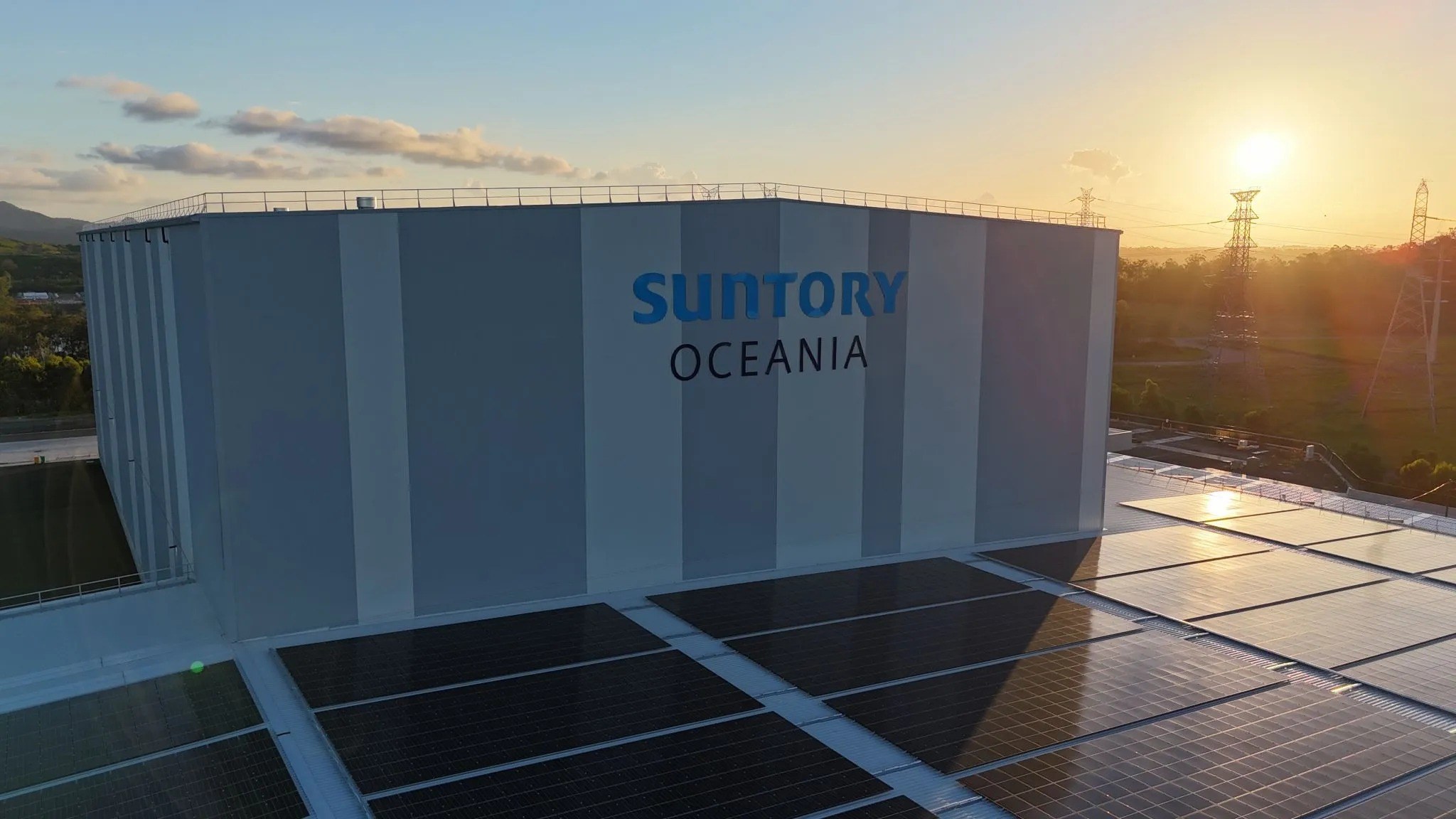 Suntory Oceania facility with solar panels at sunset, featuring large cylindrical storage tanks and company branding on white walls.