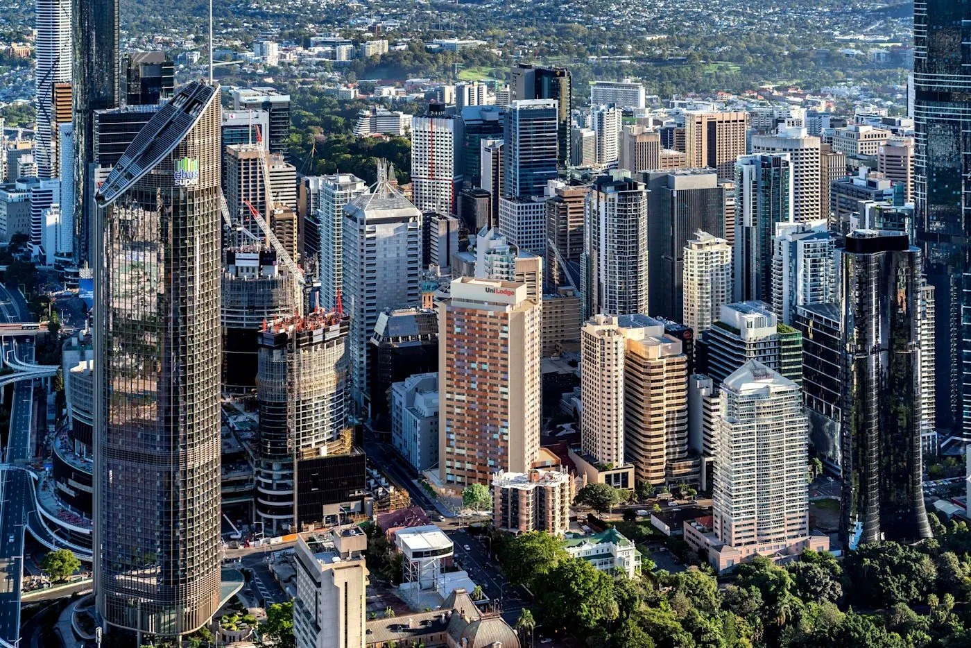 Aerial view of Brisbane city skyline with high-rise buildings, green suburban areas, and modern skyscrapers under blue sky.